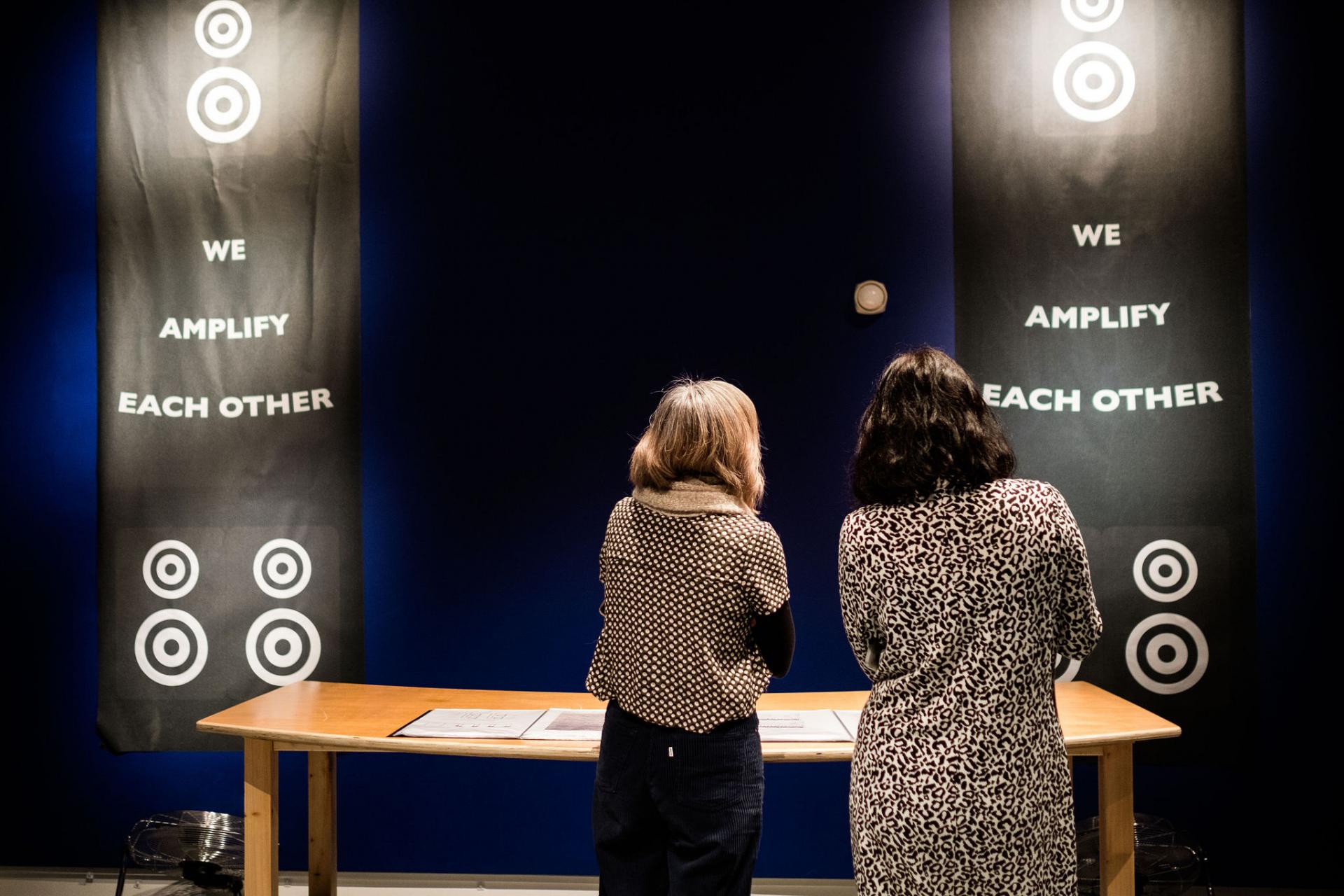 two women look at a banner with the words 'we amplify each other' on it