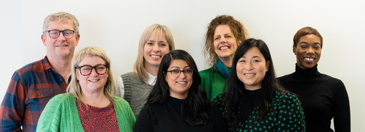 Seven people stand in front of a white background, smiling