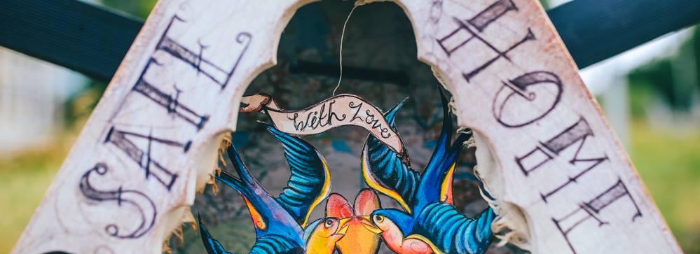 A handcrafted bird box with the words safe home written on the arches. A delicate tattoo inspired bird motif, brightly coloured bridges the gap in the box opening in front of a bottle with a ship in it.