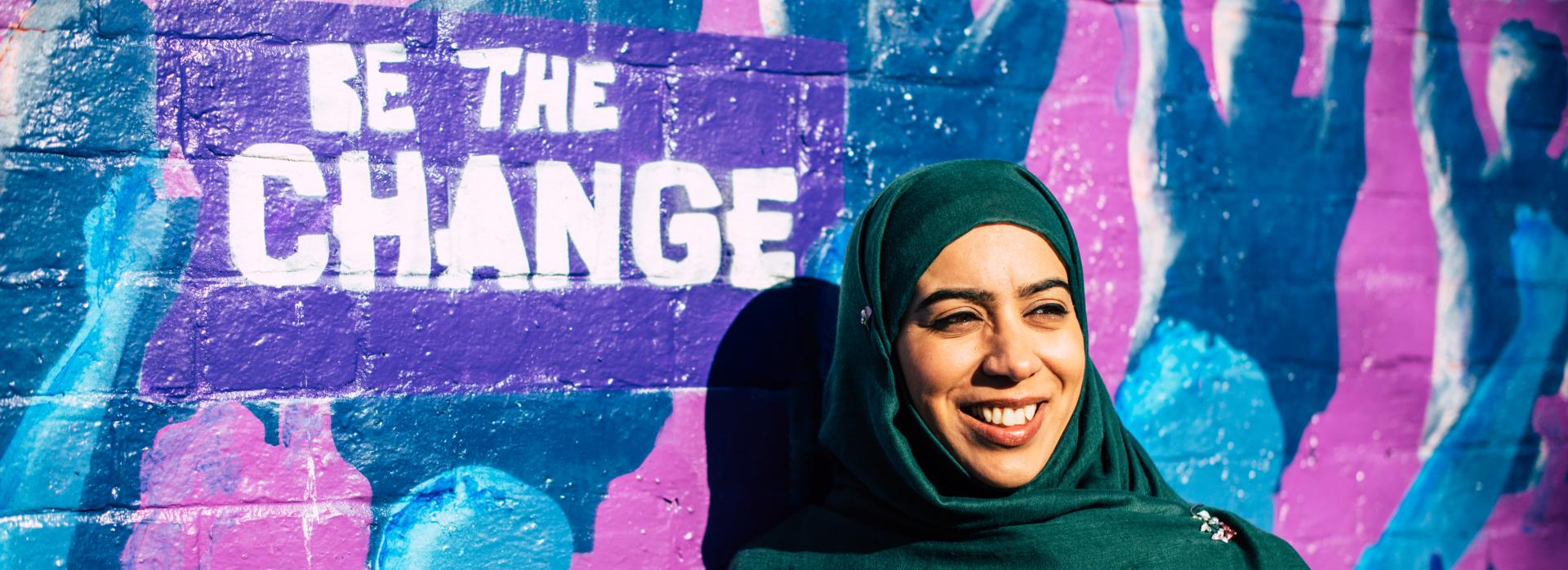 A South Asian woman wearing a dark green coloured headscarf, smiling slightly away from the camera, standing in front of a colourful graffiti wall mural with the words "Be the change" 