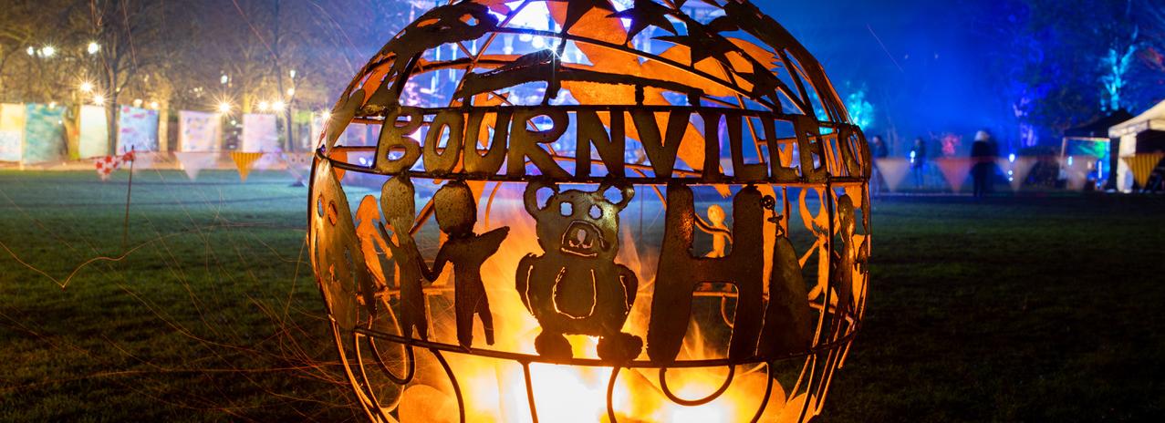 photo of a field with people in the distance at night. In the foreground is a large spherical sculpture with the word bourneville running around it above some figures, all cut out of metal; inside the sculpture is a light source