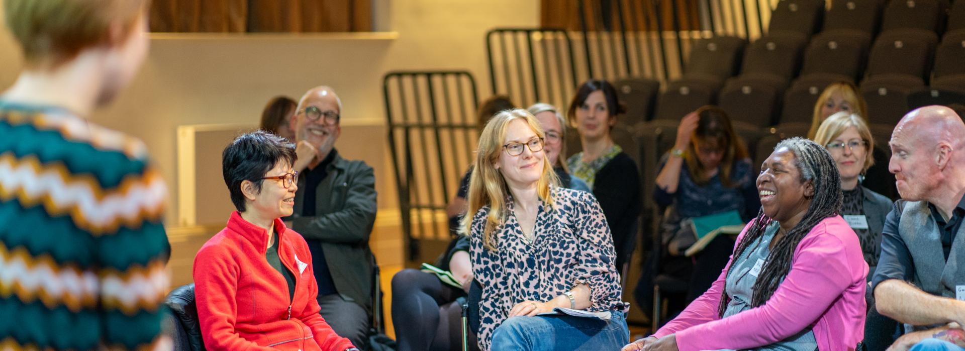 A white woman wearing glasses with short black hair and a red top sitting next to a white woman with shoulder length blonde hair who is sitting next to a black woman with locs wearing a pink cardigan smiling