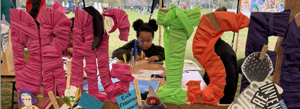   	 A portrait image. A pink fabric banner that reads 'Drag Declares Emergency Glean Hope' in green fabric letters. Paper mythical beasts surround the banner, created by workshop attendees. The banner is attached to a wooden sign that reads 'Drag is Folk' 