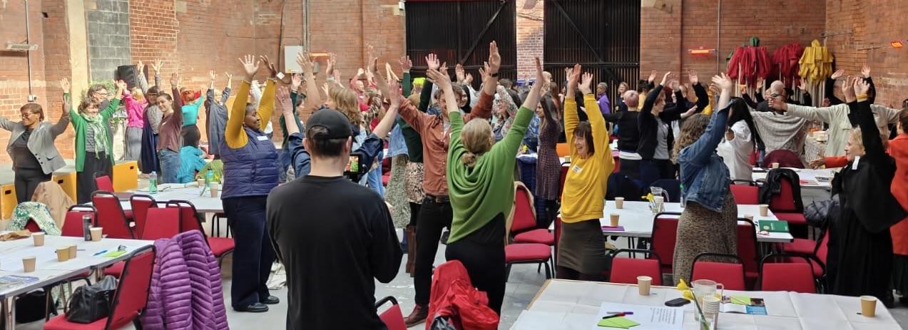 group of people in a large room wearing varying brightly coloured clothing standing with their hands in the air surrounded by tables and chairs