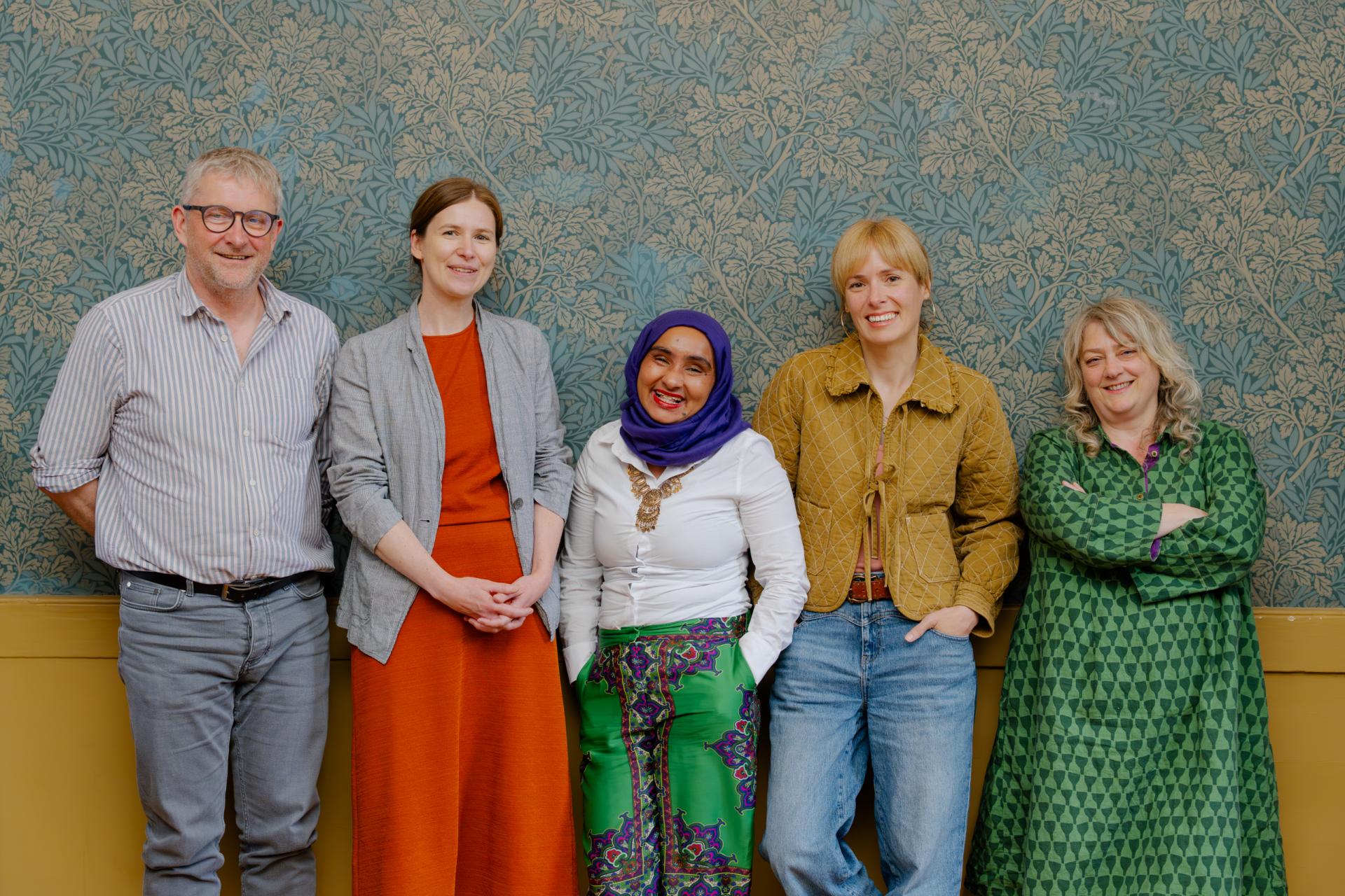 a group of people wearing colourful clothing smiling at the camera, standing against blue and gold printed wallpaper