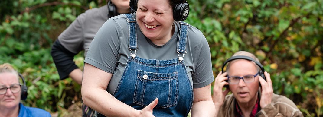 four adults stand and sit with headphones on, the woman in the foreground is laughing as she listens