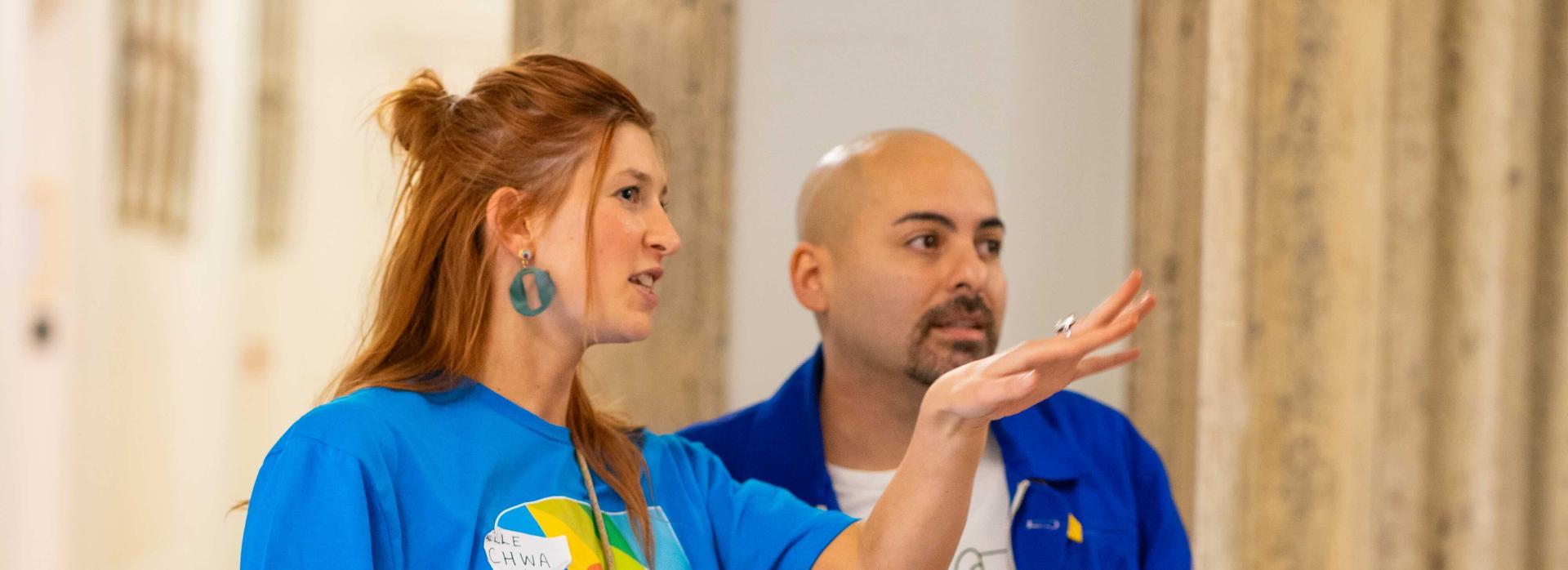 A woman with light brown wearing a blue t-shirt and a black skirt directing a bald headed male wearing a blue shirt over a white t-shirt and red trousers