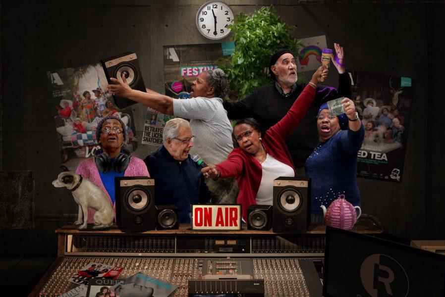 image shows a group of older people in a radio studio posing with dramatic expressions. There are speakers and a mixing table, a clock showing 11.30am and some posters on the back wall. 