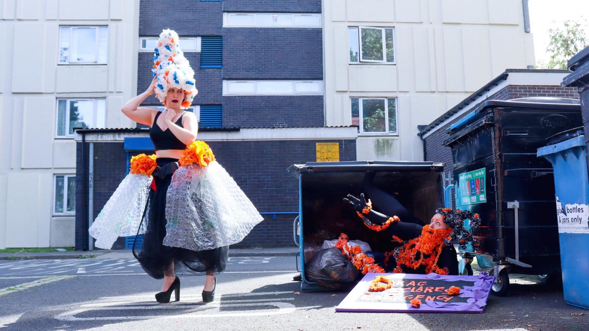 Colour photo, exterior. Kitt, a shaven headed white human, is crawling out of an industrial sized bin with arms outstretched towards Sarah a white human who is looking appalled. They are both wearing extravagant drag outfits made from garlands of flowers handmade from recycled black, orange, white and blue plastic bags.