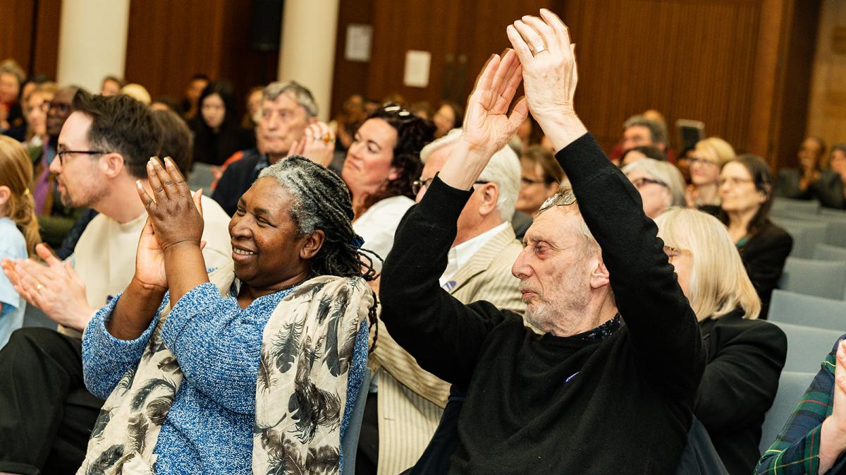 People clapping enthusiatically in an audience at an event - one clapping their hands over their head 