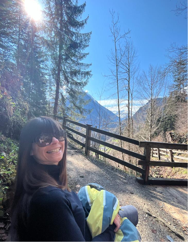 A person sitting on a bench smiling, with a fence, mountains and blue skies in the background.