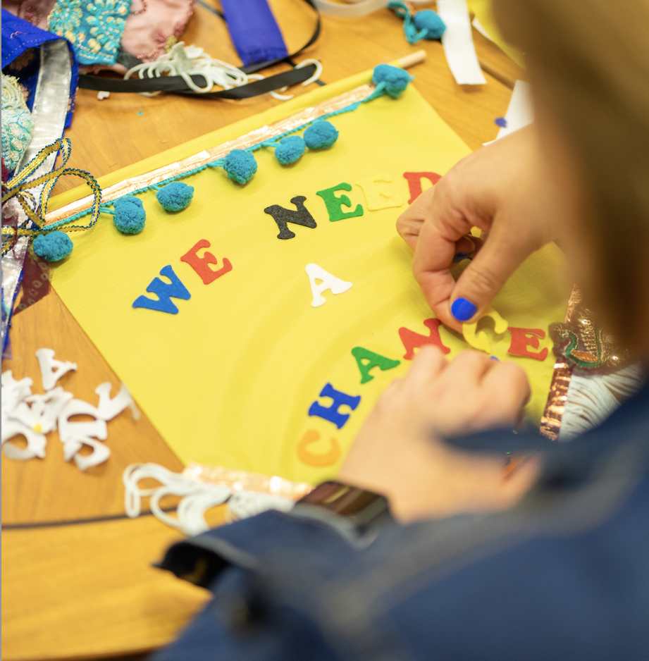 A person making a colourful banner that reads 'We Need A Change'