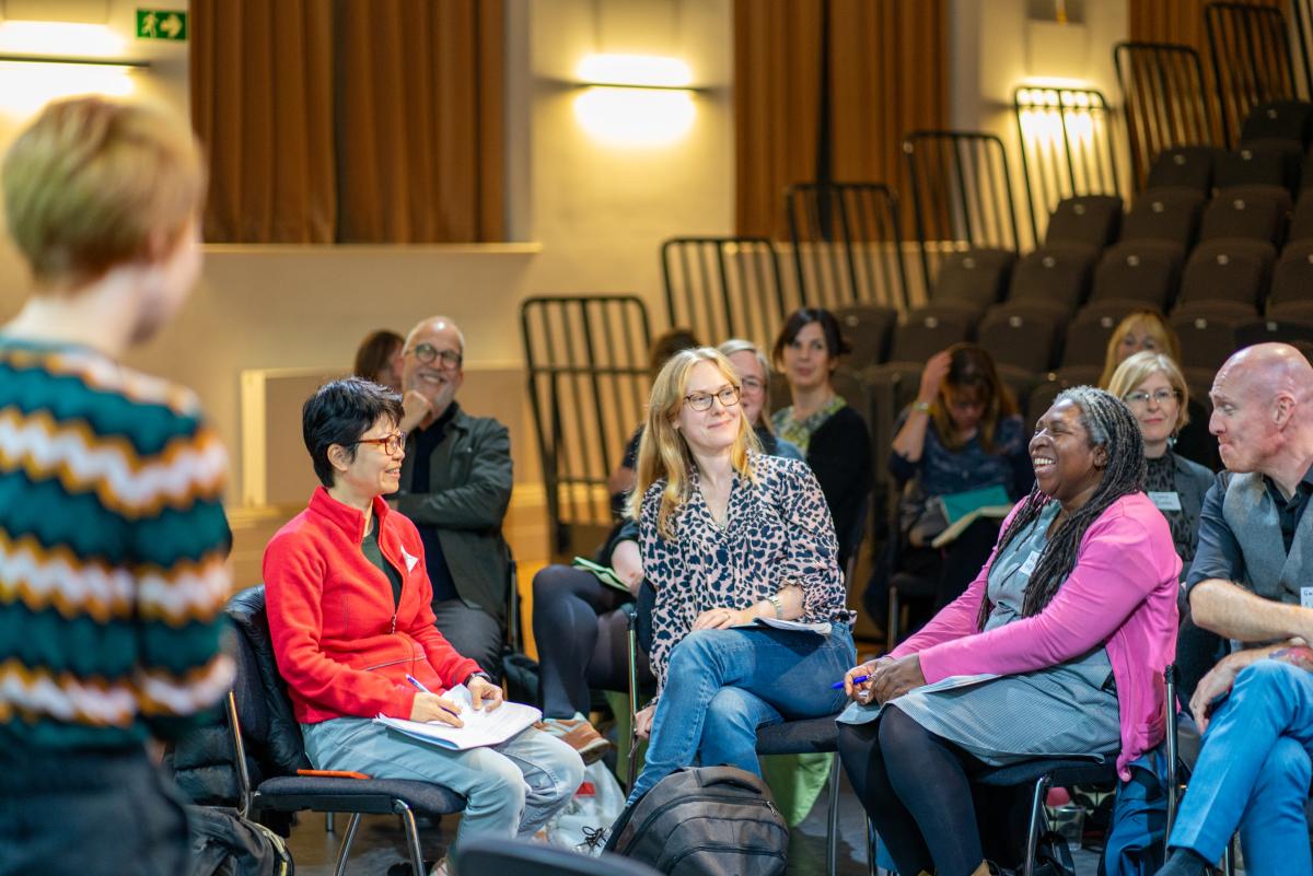 A white woman wearing glasses with short black hair and a red top sitting next to a white woman with shoulder length blonde hair who is sitting next to a black woman with locs wearing a pink cardigan smiling
