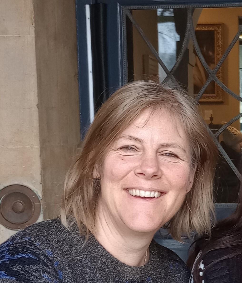 close-up photo of a person with medium-length blond hair smiling ino the camera with a door frame behind them