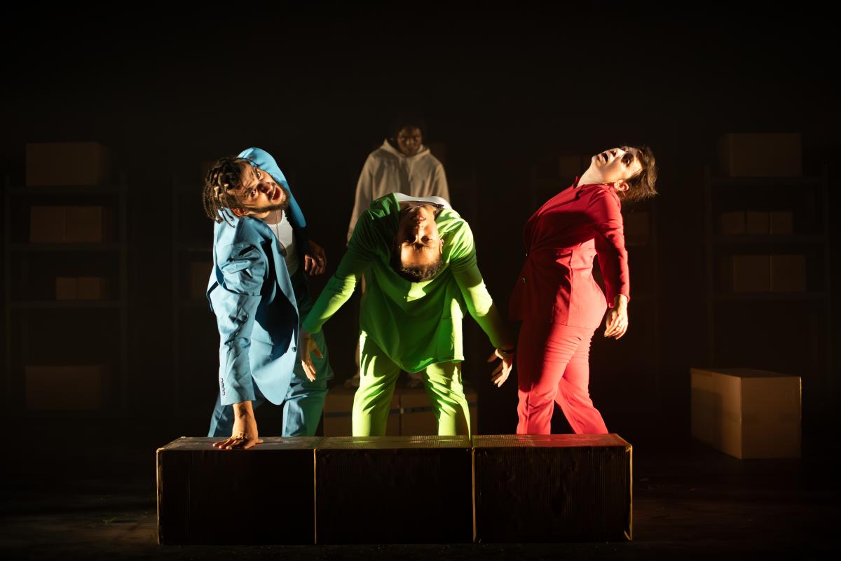 three people in brightly coloured suits stand close to each other on a stage with a black background, all leaning at unusual angles