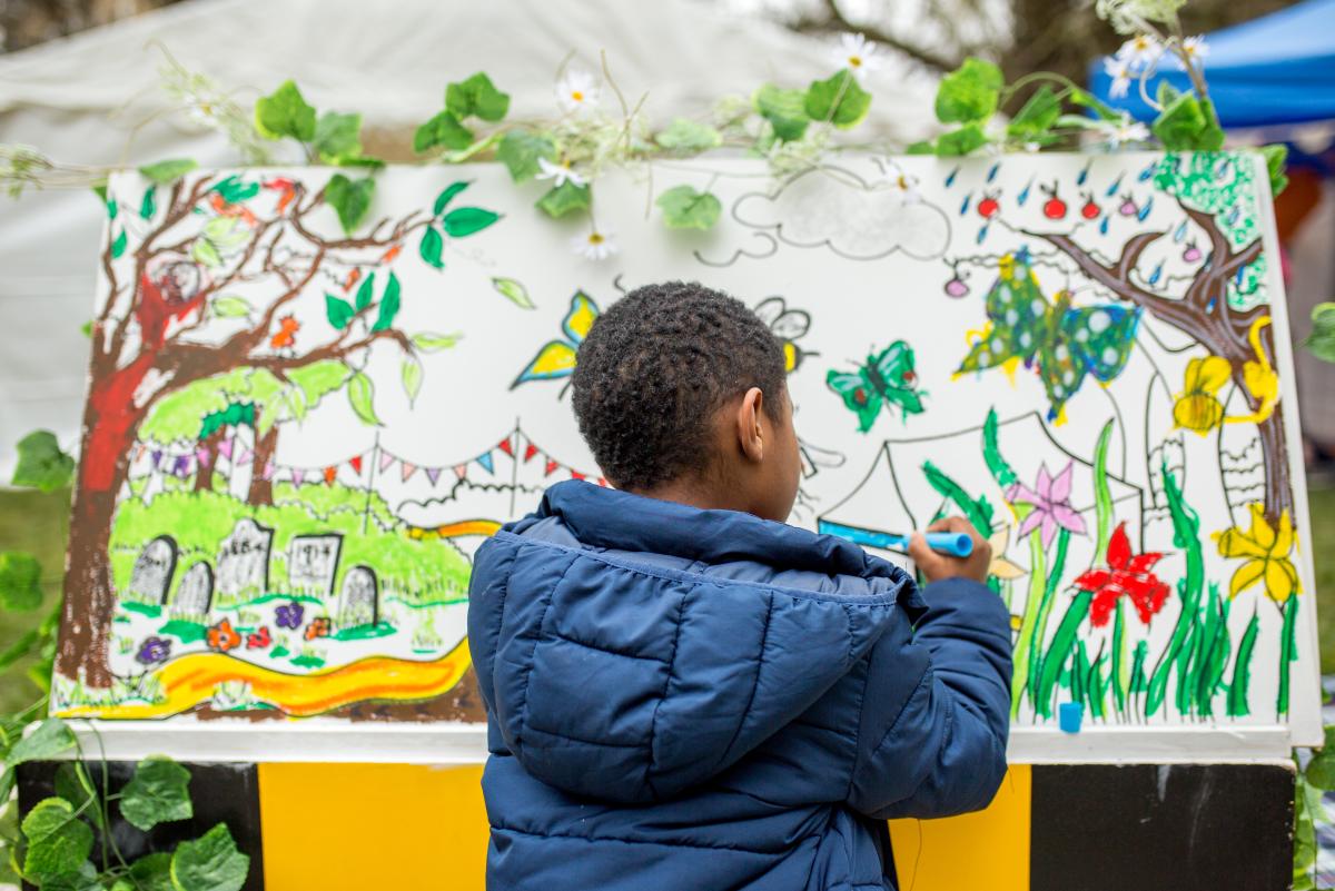Young black boy wearing a navy coat, back to the camera drawing on a colourful canvas