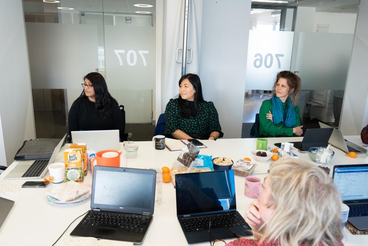 four people sit around a table covered with laptops, snacks and papers