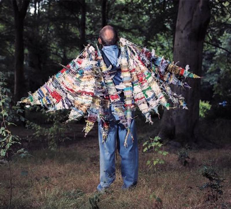 A man facing away from the camera and into a wood. On his back are large wings made of scraps of fabric.