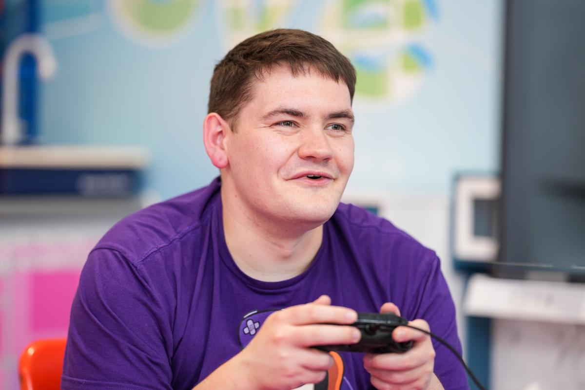 Young man with black short hair, wearing a purple t-shirt holding a console control pad and smirking