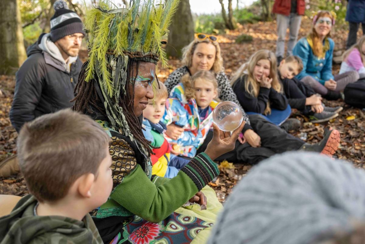 A woman holds up a glass ball, sitting in a circle surrounded by young people and their families in a natural setting