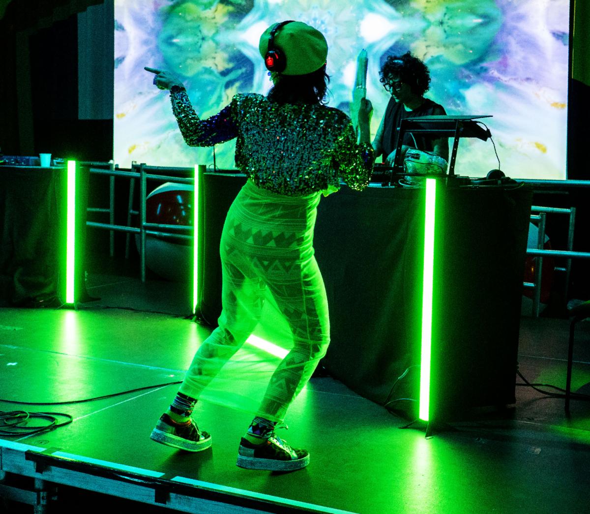woman dancing with her back to the camera wearing headphones under a green hue with a dj on the decks in the background