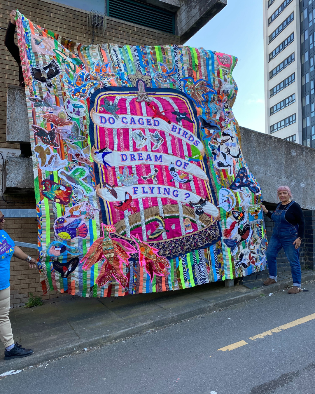 A woman wearing blue dungarees with blonde hair holding a large colourful quilt by the roadside