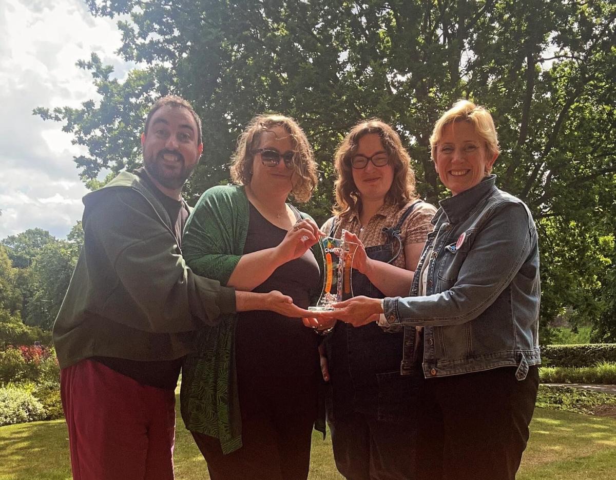 Four people, the leadership team of Next Door But One, standing in the sun in front of trees looking happy, all holding a bright and colourful glass award with ‘CHWA -  Culture Health and Wellbeing Alliance’ on it