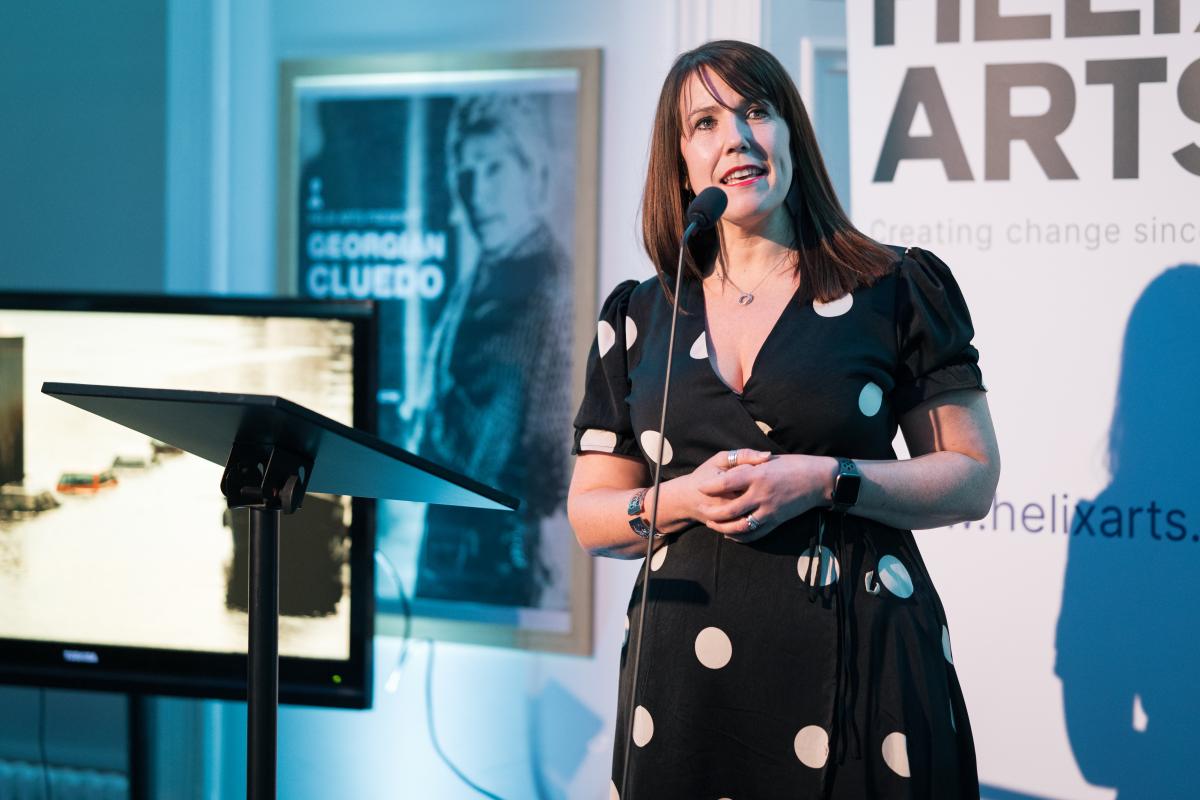 A white woman with shoulder length brown hair wearing a black dress with white circles, standing in front of a microphone speaking against a white background with black text