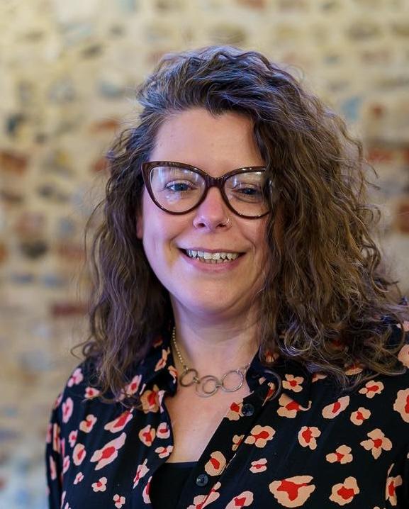 A white woman with shoulder length brown curly hair wearing glasses wearing a black flowered shirt smiling  against a brick background