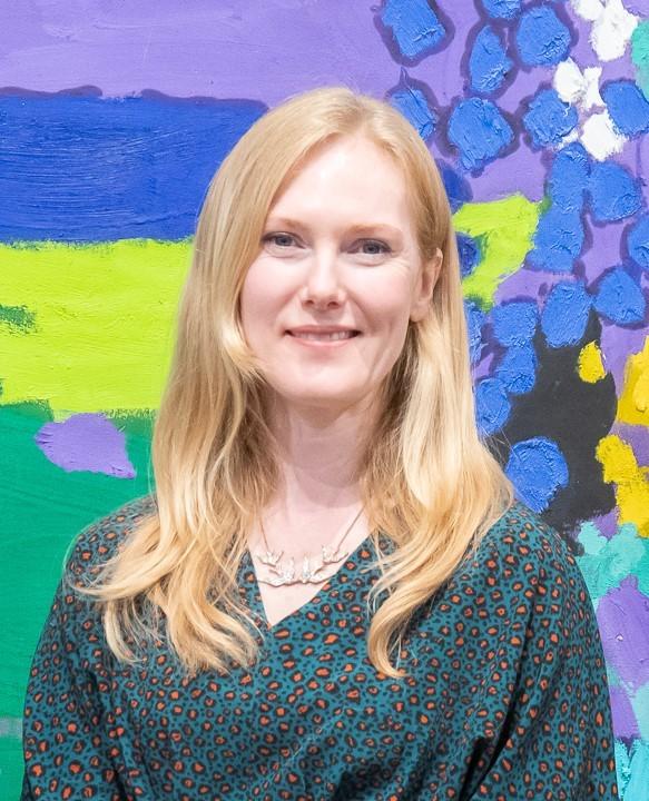 A white woman with shoulder length blonde hair wearing a green v neck dress smiling at the camera against a multi-coloured painted background
