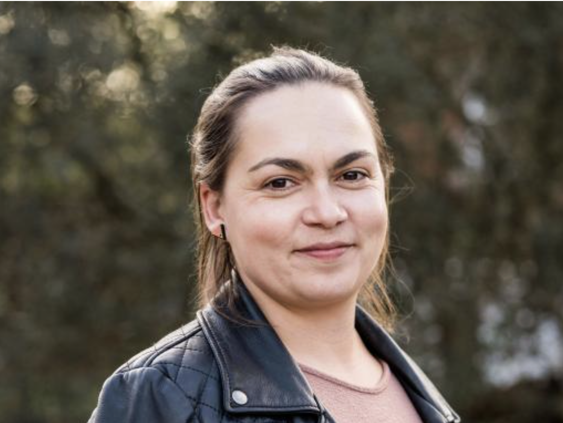 A white woman with dark brown haired tied into a ponytail wearing a black leather jacket and a brown top against a background of trees