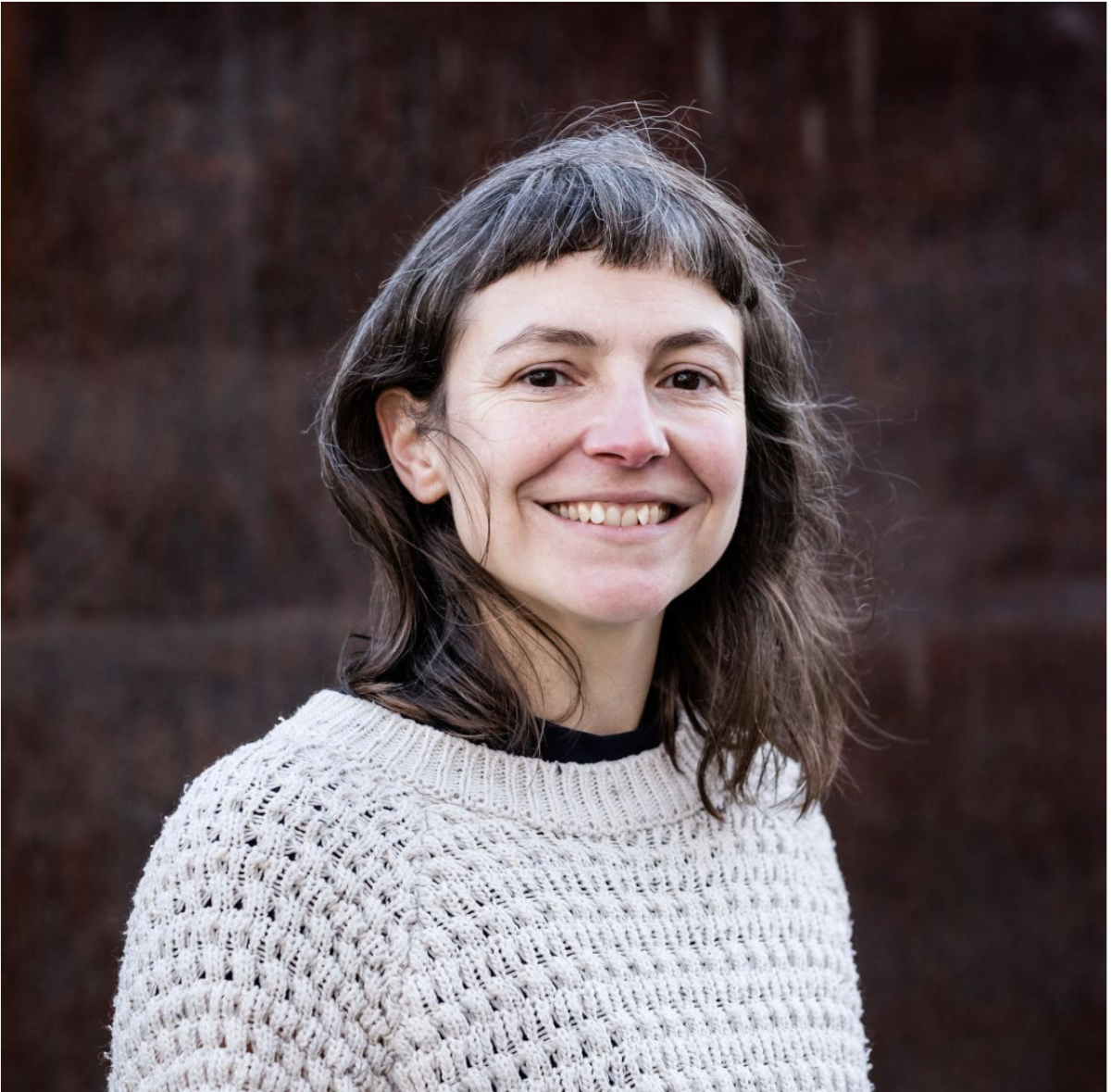 A white woman with brown shoulder length hair wearing a white wooly jumper smiling against a brown brick background
