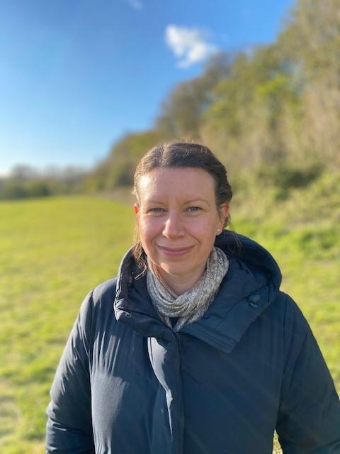 A white woman with her hair tied back wearing a black puffer jacket and a grey scarf against a nature background