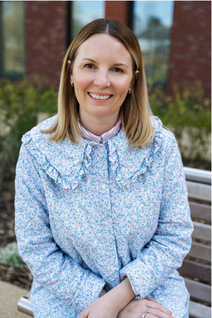 A woman with brown shoulder length hair wearing a blue frilly dress with her hands in her lap smiling at the camera