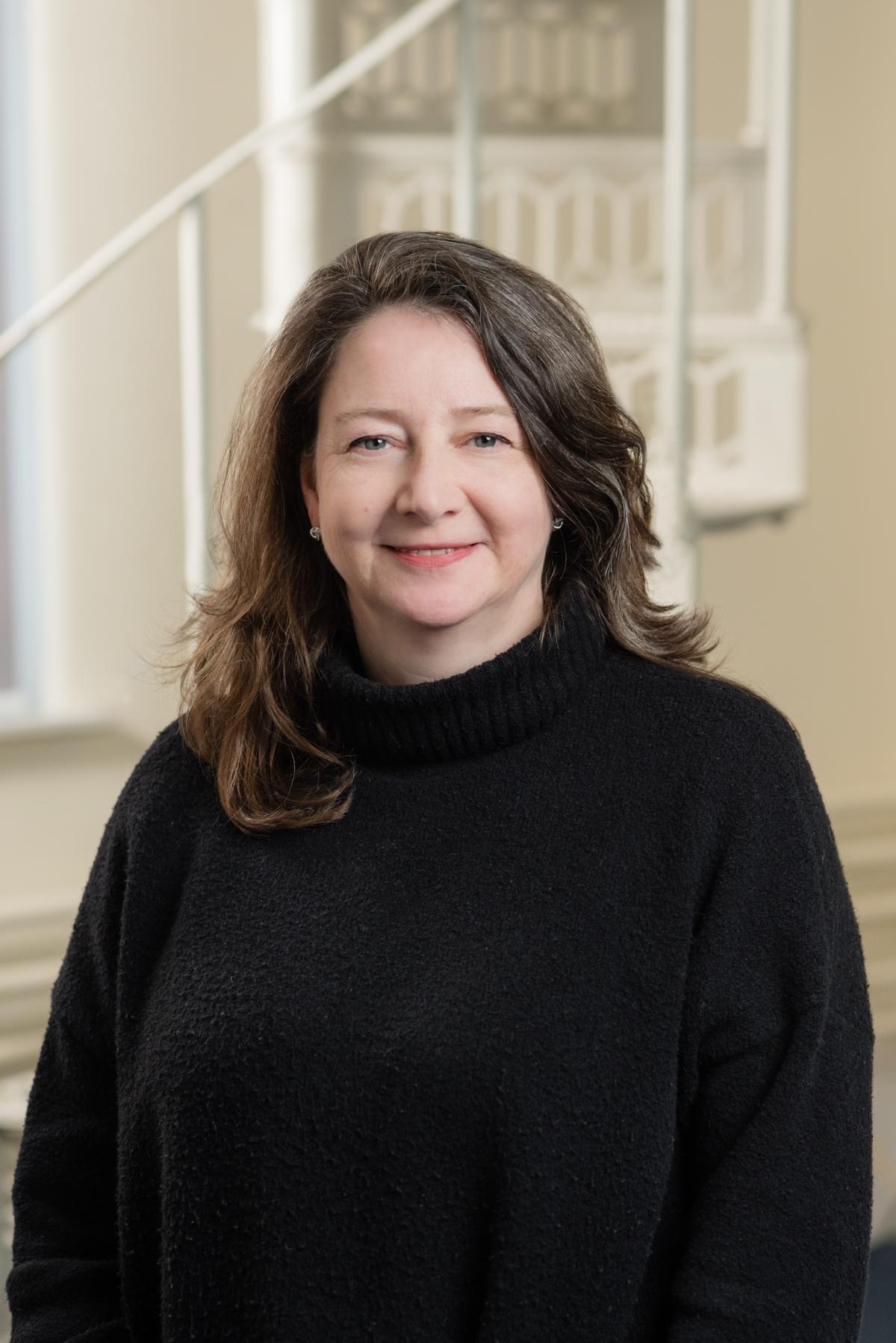 Half-length portrait photo of a woman with long dark hair and a black jumper stannding in front of white bannisters
