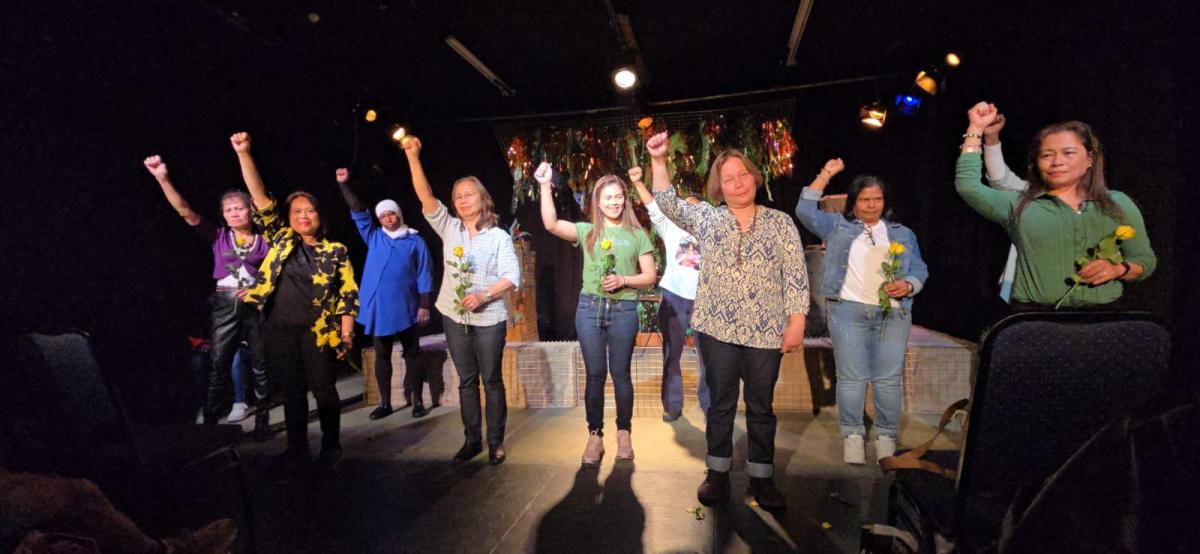 A group of people wearing multi-coloured clothing standing on a stage with dim lighting standing with their hands in the air