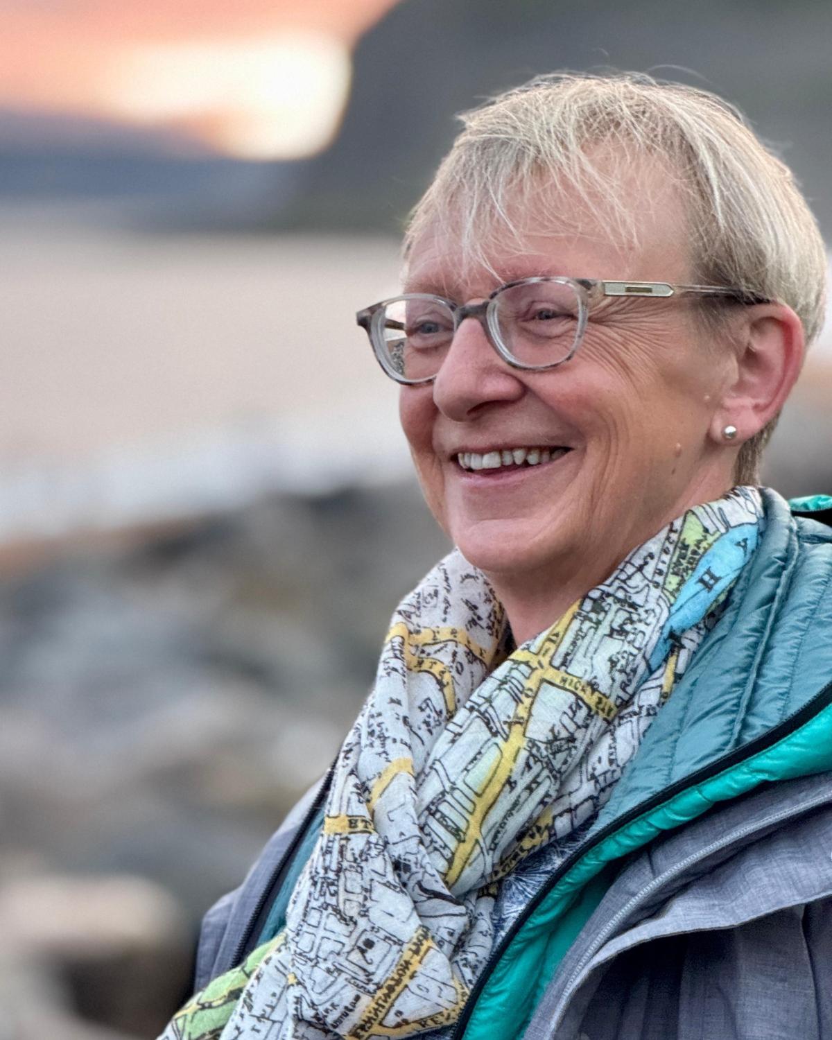 portrait of a woman with short blonde hair and glasses smiling, on a beach