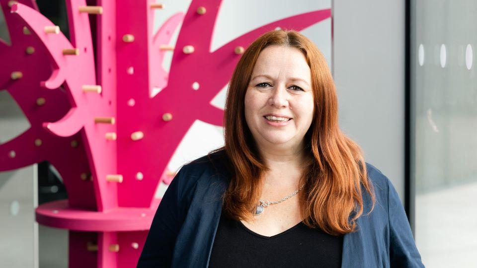 A white woman with mousy brown shoulder length hair smiling at the camera against a pink painted background