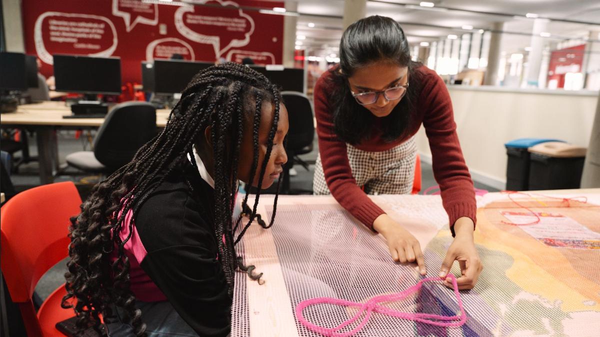 two children leaning over a desk creating a piece of art work with pink thread and coloured paper