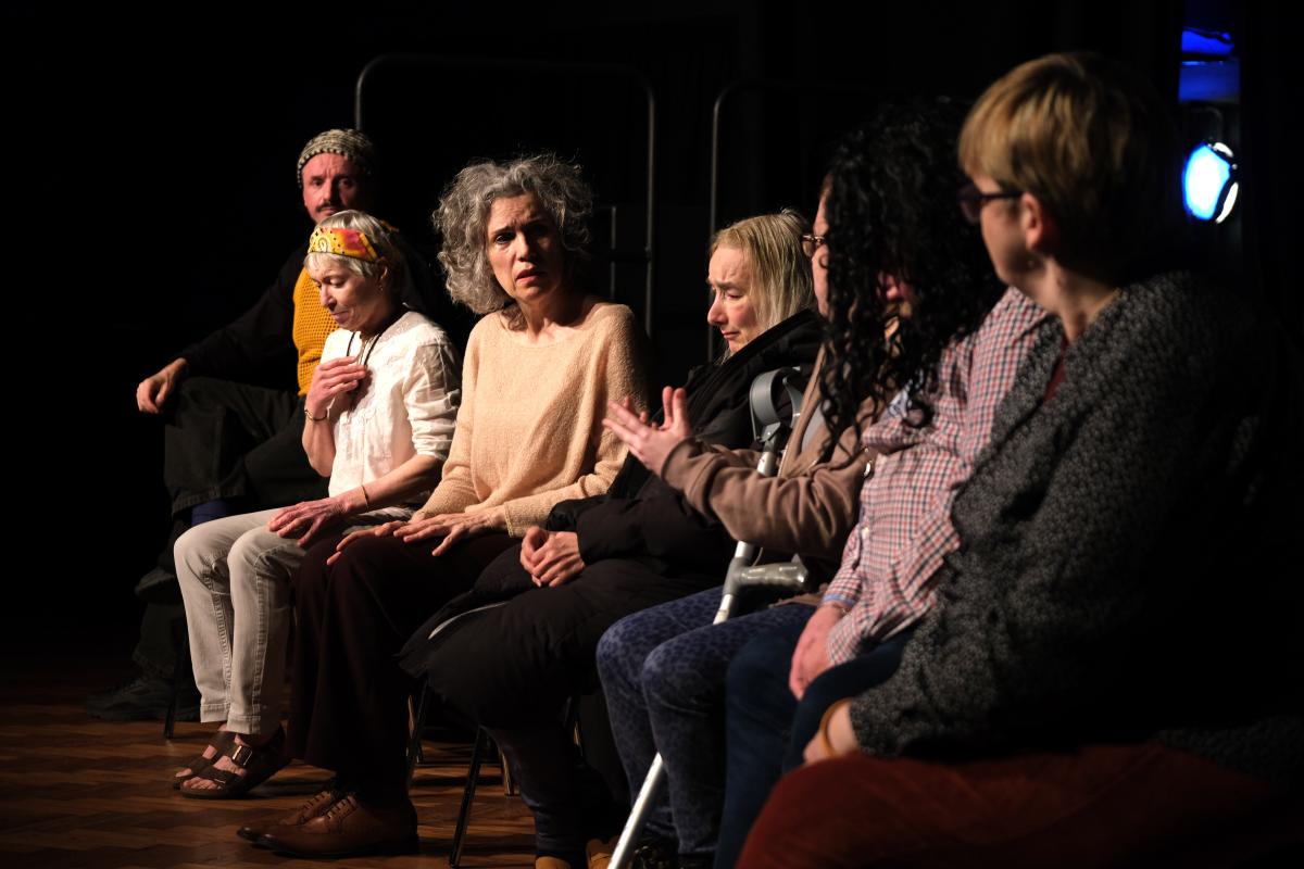 A group of people sitting down side by side upon a dimly lit stage conversing with one another