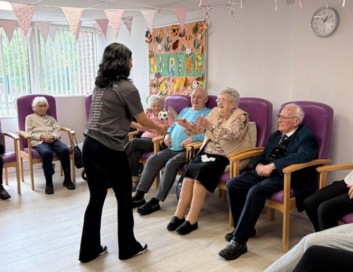 A woman with long black hair wearing a grey t-shirt and black trousers standing and gesturing to older people sitting in purple chairs facing her