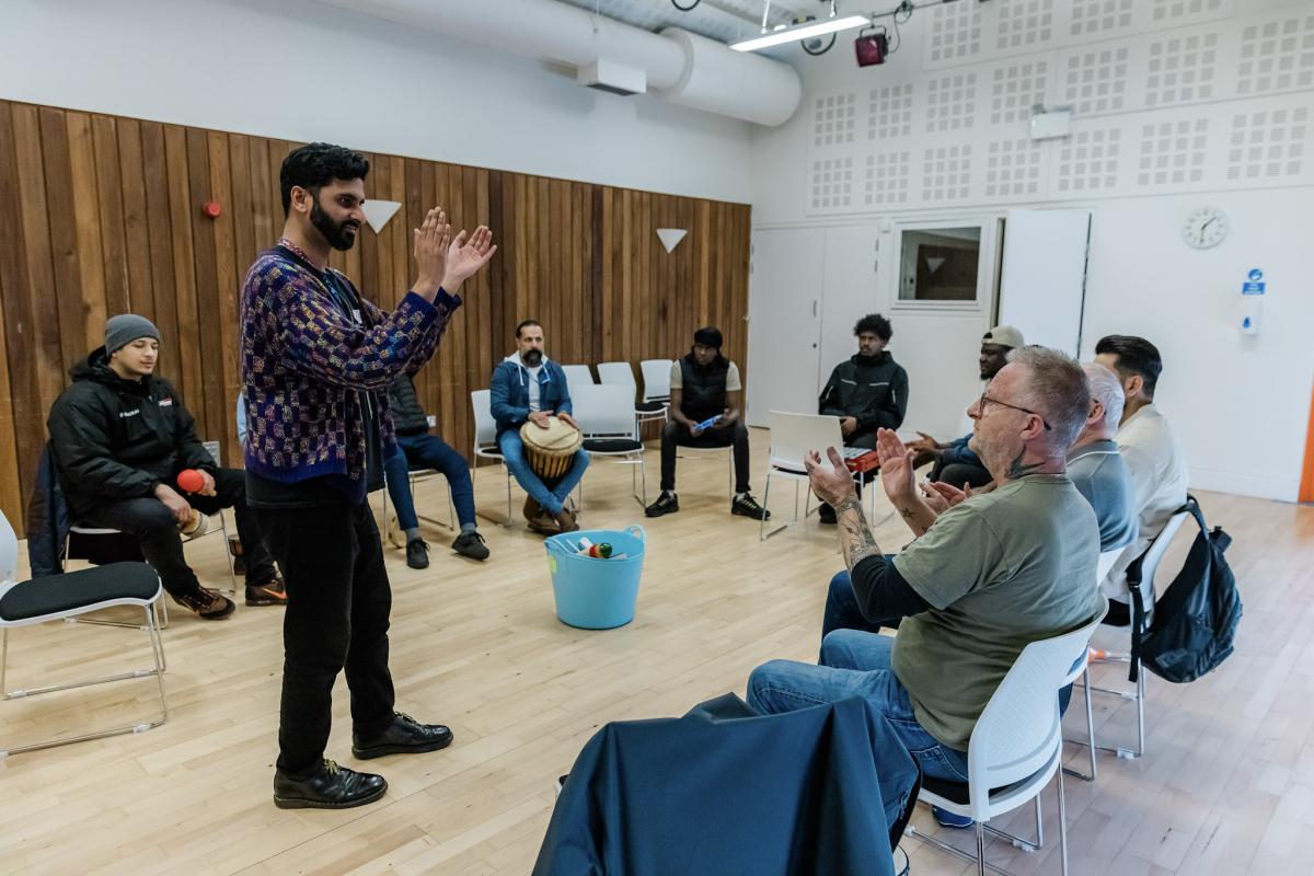 A bearded male with short black hair wearing a dark coloured wooly jumper with flecks of colour and black jeans standing in front of seated individuals in a large community space with his hands held in a clapping stance