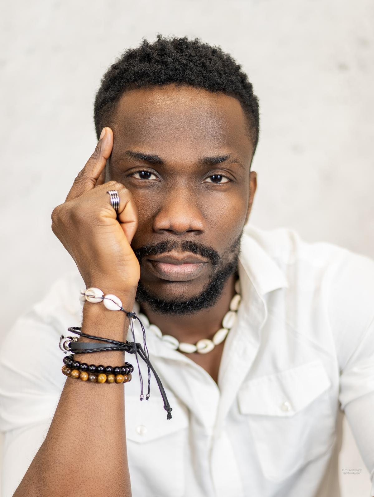 A black man with his hand to his head wearing bracelets and a white shirt against a white background