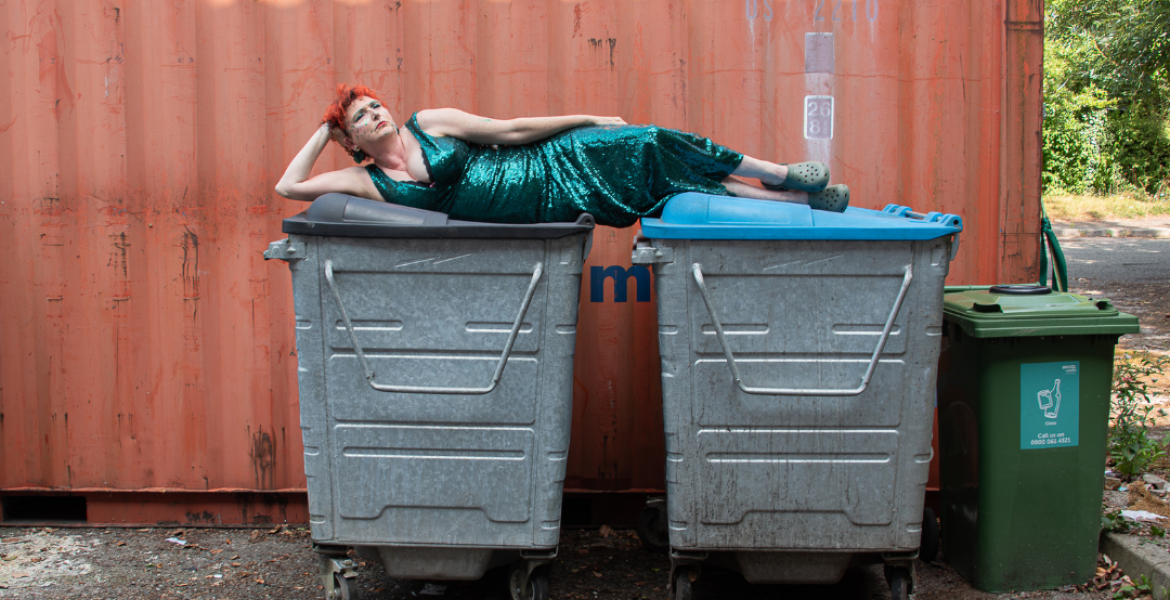 a woman with red hair wearing a green sparkly dress lying across two bins against a red steel background
