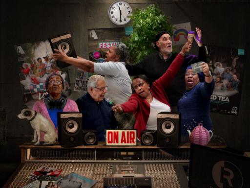 image shows a group of older people in a radio studio posing with dramatic expressions. There are speakers and a mixing table, a clock showing 11.30am and some posters on the back wall. 