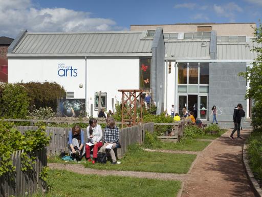 People gather in a garden with a path leading to a white-painted building with 'North Edinburgh Arts' written on the side of it