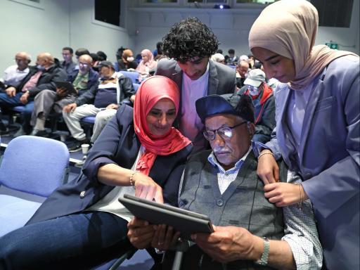 Members of the Connecting with Yemeni Elders' Heritage looking together at the app created on an iPad in a theatre of people