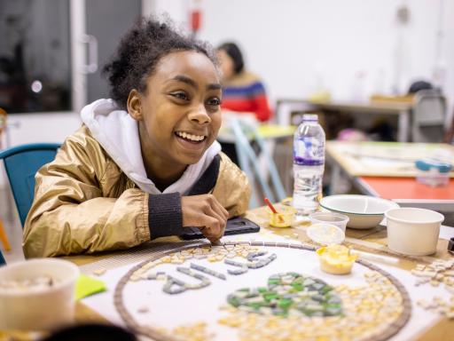 Young girl sits at a table making a mosaic and smiling