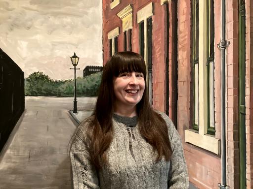 Joanne Charlton, North East Regional Champion, wearing a grey jacket stood in front of a painted scene of a victorian street