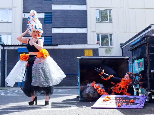 Colour photo, exterior. Kitt, a shaven headed white human, is crawling out of an industrial sized bin with arms outstretched towards Sarah a white human who is looking appalled.  They are both wearing extravagant drag outfits made from garlands of flowers handmade from recycled black, orange, white and blue plastic bags. 